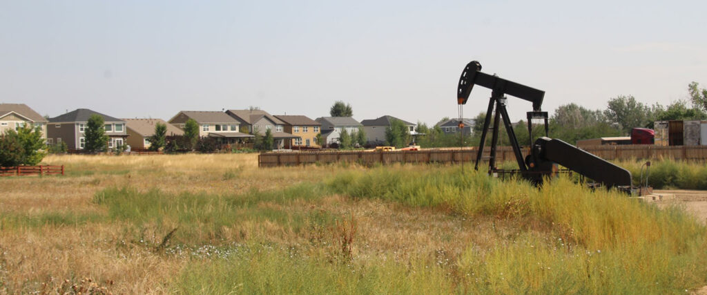 Dirt road with weeds and an oil derrick to the side of a green meadow with suburban homes in the background